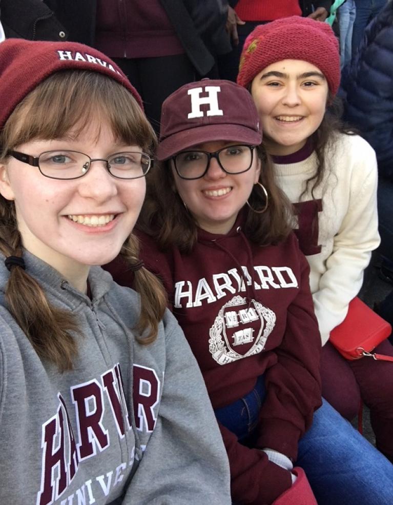 Three girls in Harvard gear take a selfie in the bleachers of a football stadium.