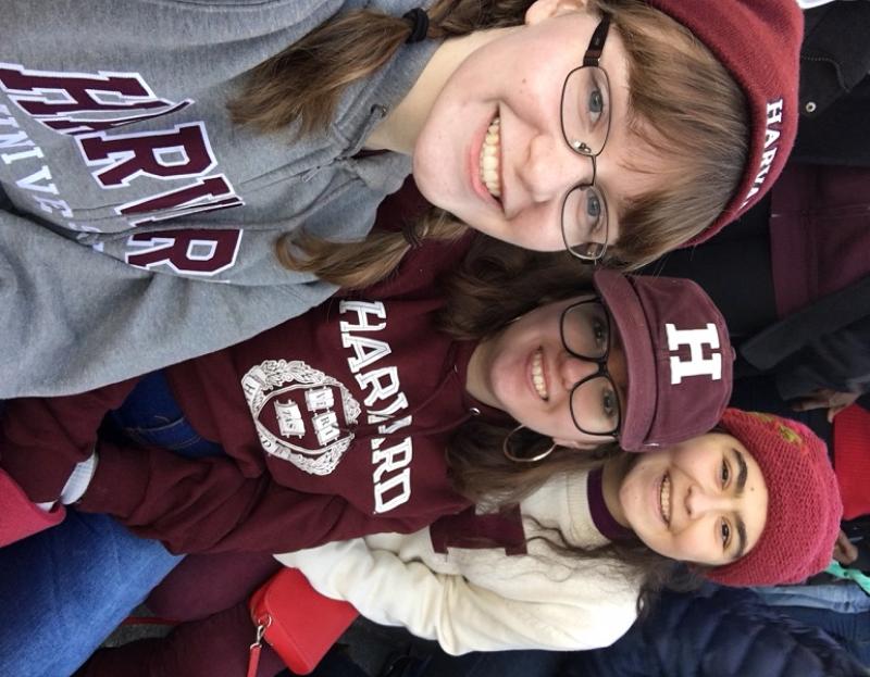 Three girls in Harvard gear take a selfie in the bleachers of a football stadium.