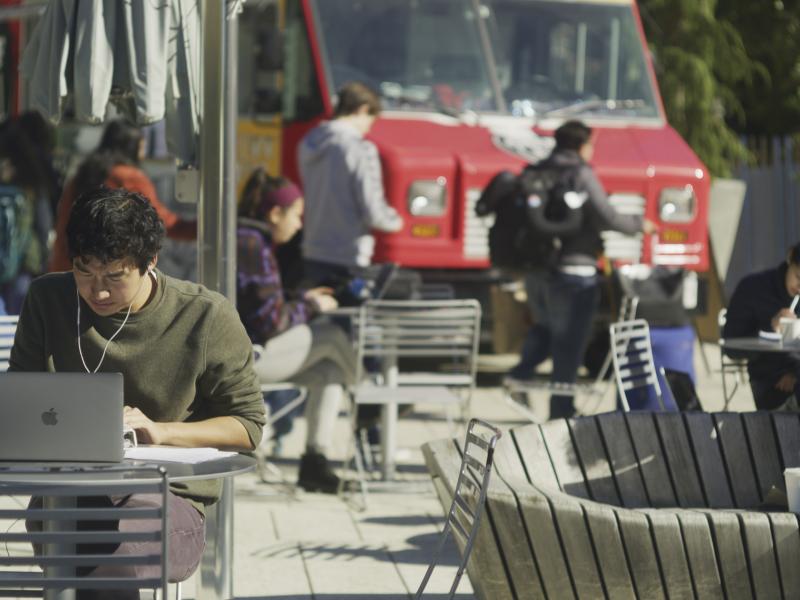 A student in a green sweater sits at a laptop at an outside table. Students in the background are getting lunch from a foodtruck.