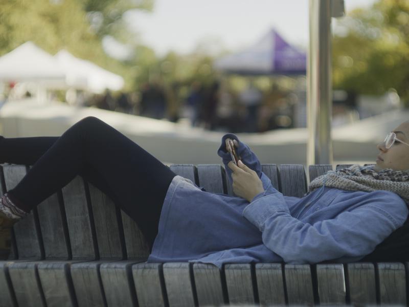 Student laying on bench at Science Center Plaza with her phone