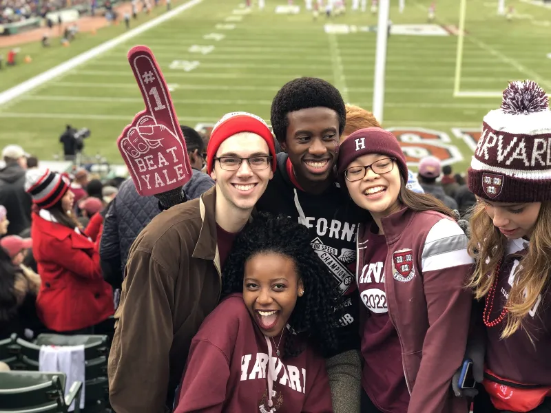 Four students cheering in the stands at a Harvard football game