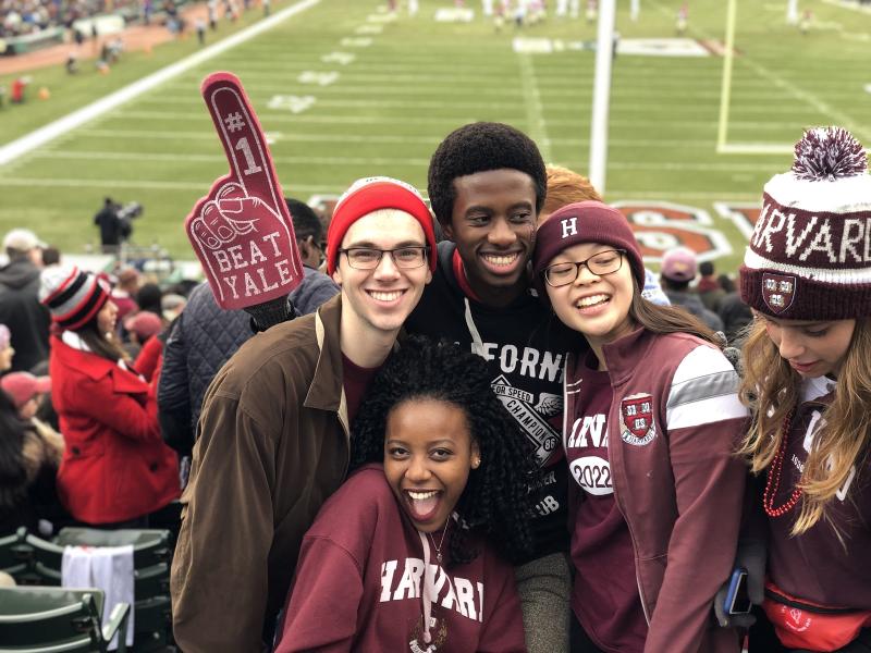 Four students cheering in the stands at a Harvard football game