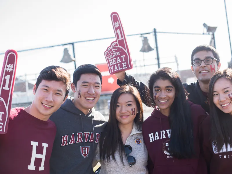 Six Harvard students with foam fingers cheering for Harvard-Yale football game