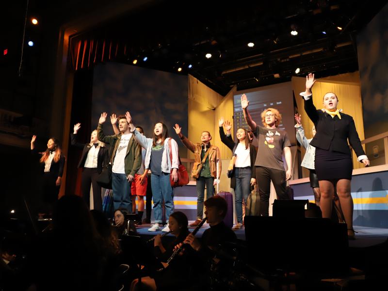 Actors on stage raising their hands singing, with musicians below