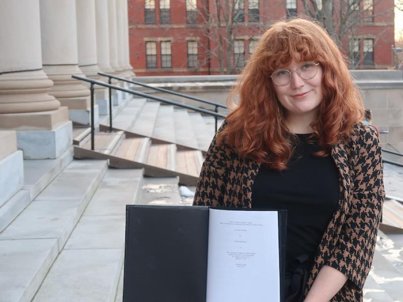 Allison standing on the steps of a large library holding a black thesis binder opened to the title page of her thesis.