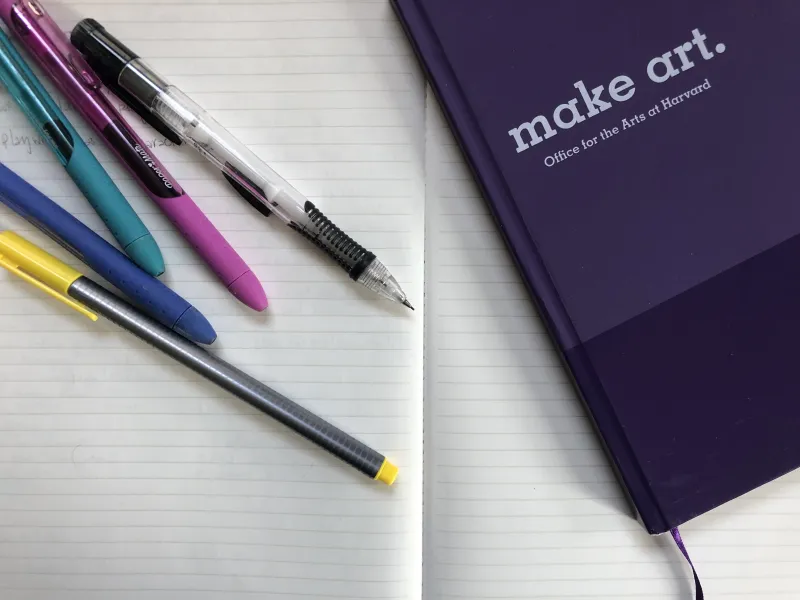 An open notebook. On top there is another purple notebook that says "Make Art" on the cover, along with a variety of pens and pencils.