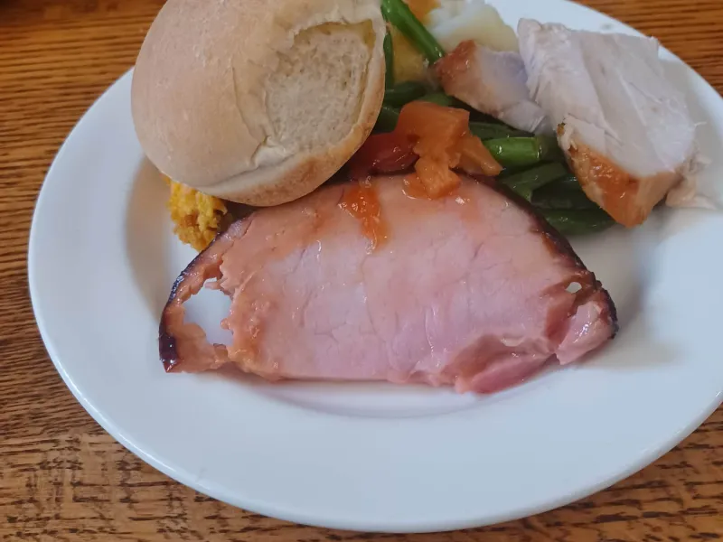 White plate piled with a slice of ham, turkey, a bread roll, green beans, and sweet yams.