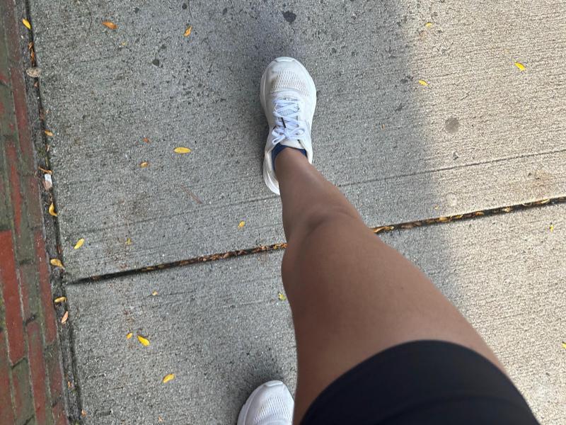 A girl's tan leg wearing black running shorts and white running sneakers, mid stride on a concrete sidewalk.