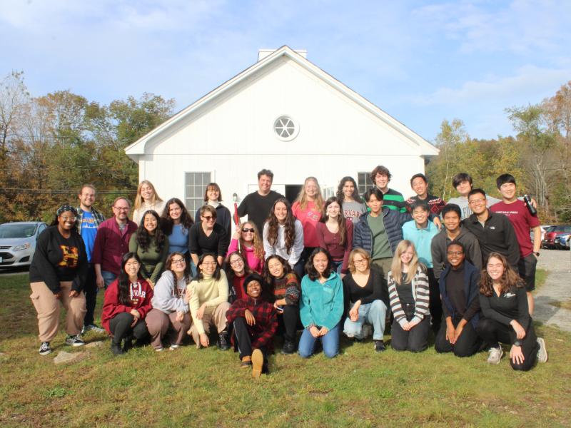 Picture of the Harvard-Radcliffe Collegium Musicum standing in front of a white cabin under blue skies on a green hill.