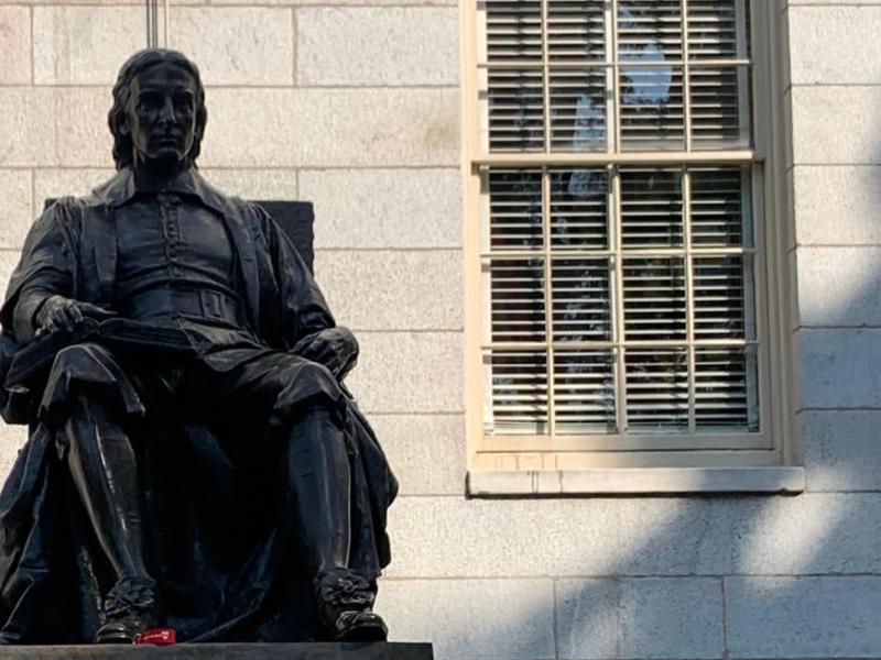 This is a picture of the top of the John Harvard Statue, which shows only the shape of the statue itself, and not the base with it's description. The statue shows a man posing in a sitting, very academic looking, position. 