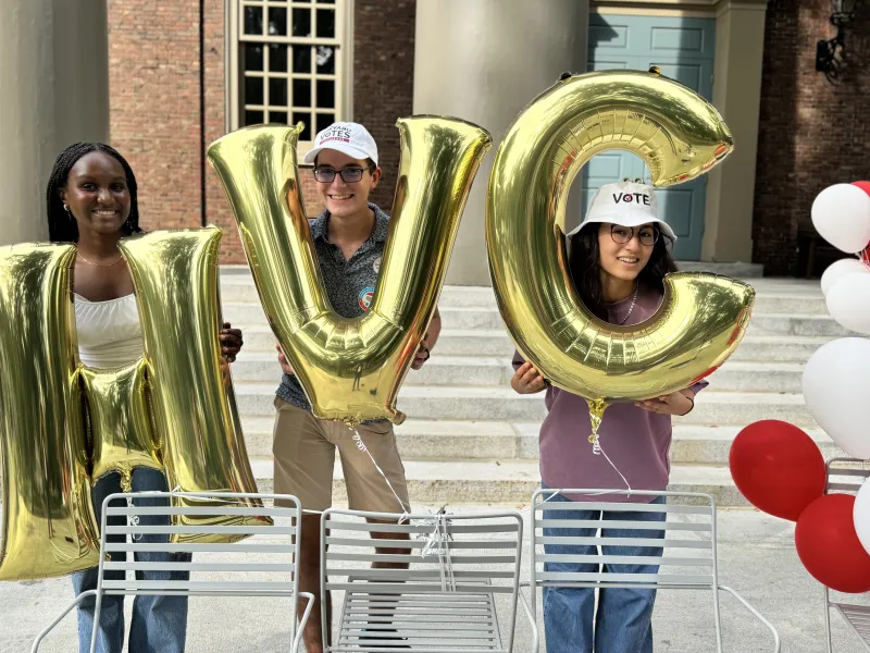 Three students holding large gold balloons forming "HVC" (Harvard Votes Challenge). They wear "VOTE" hats, situated in front of a campus building with white and red balloons nearby, smiling in a lively, outdoor setting.