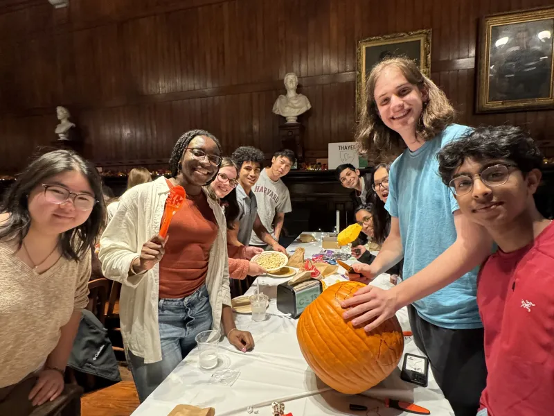 Students holding pumpkin-carving tools stand around a table in Annenberg Hall. On the table sits a pumpkin and several pieces of candy.