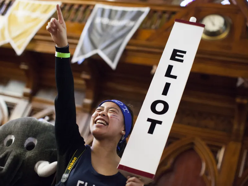 A person smiling and cheering, holding an "ELIOT" sign, with banners and a wooden interior background.