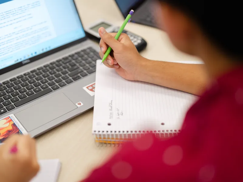 A person is taking notes in a spiral notebook with a green pencil while looking at a laptop screen