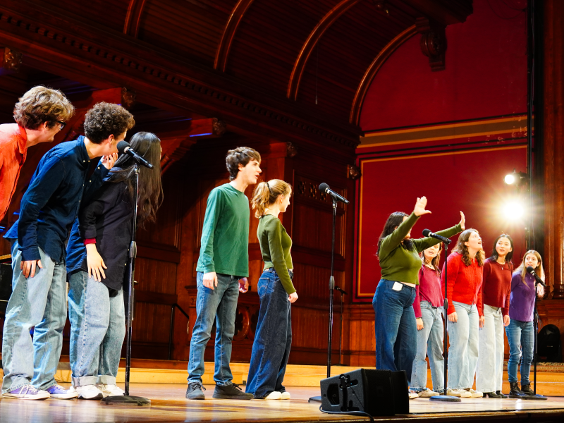 A group of performers on stage, singing into microphones, with a spotlight illuminating them.
