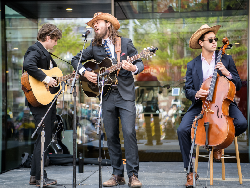 Three musicians in suits and cowboy hats perform outdoors, playing acoustic guitar, electric guitar, and cello.