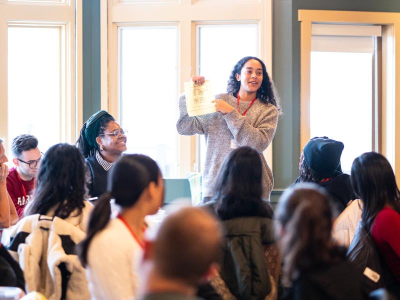 A woman stands and holds up a piece of paper while speaking to a group of people seated around her.