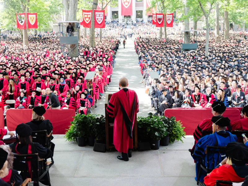 A speaker addresses graduates in caps and gowns at a large outdoor Harvard commencement ceremony.