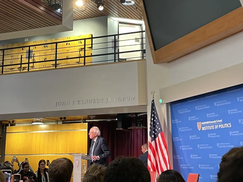 A picture of the main lobby of the Kennedy School building. It is decorated with an American flag and full of students listening to Bernie Sanders speak.