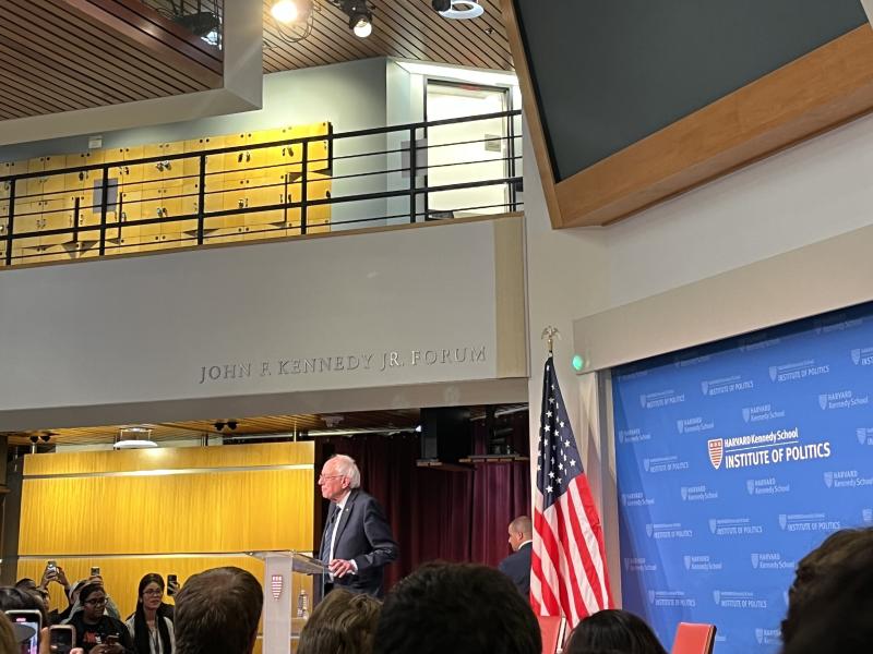 A picture of the main lobby of the Kennedy School building. It is decorated with an American flag and full of students listening to Bernie Sanders speak.