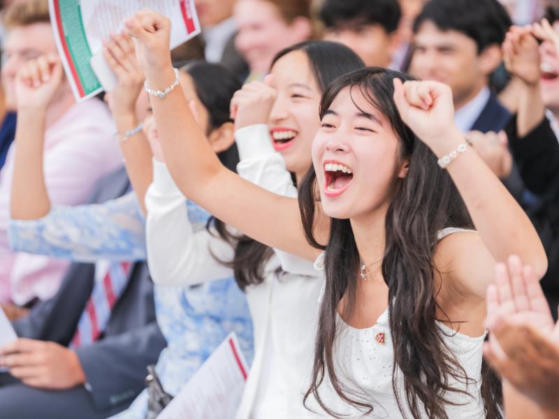 A young woman smiles cheers with delight with her hands in the air during the Convocation ceremony.