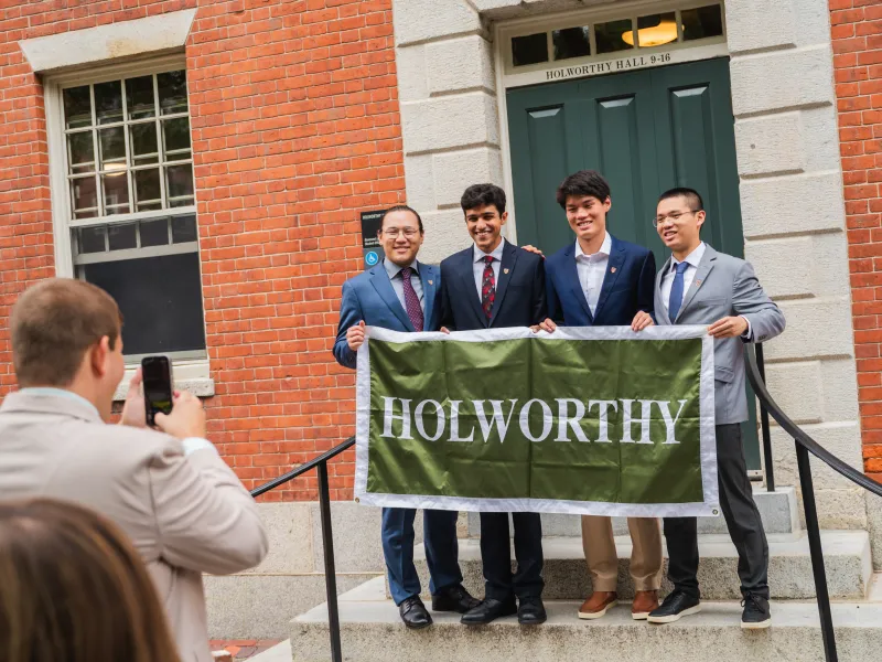 A man takes a photo of four male students of the Class of 2029 smiling for a photo together while holding a large green sign with the word "Holworthy" on it in white letters. 