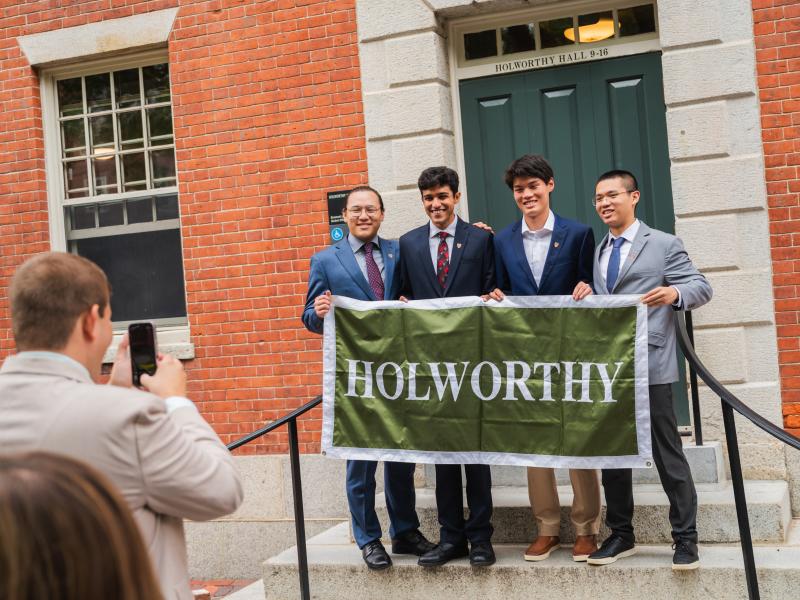 A man takes a photo of four male students of the Class of 2029 smiling for a photo together while holding a large green sign with the word "Holworthy" on it in white letters. 