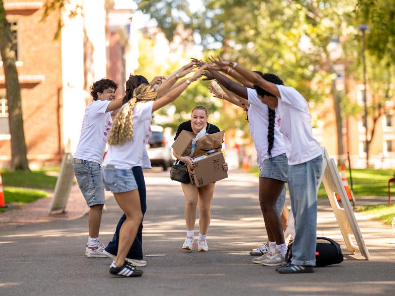 A first-year female student smiles while carrying her move-in boxes and going through a welcome tunnel made up of upperclassman students.