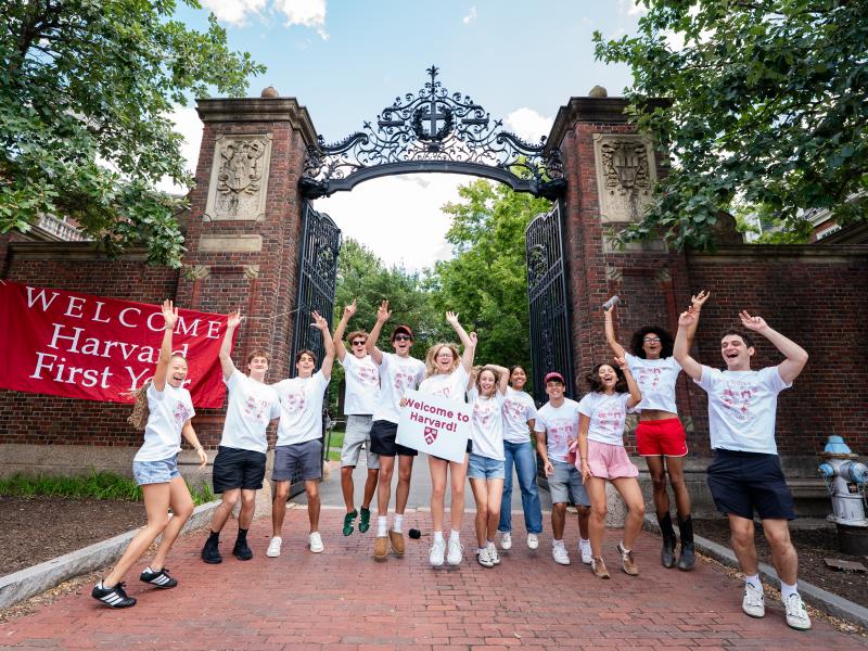 A crowd of upperclassman wearing matching white shirts cheer with a "Welcome to Harvard" sign outside of Harvard gate.