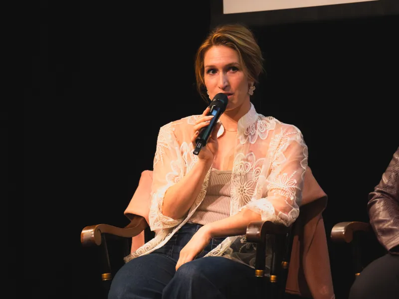 Woman sitting on a chair, speaking into a microphone on stage.