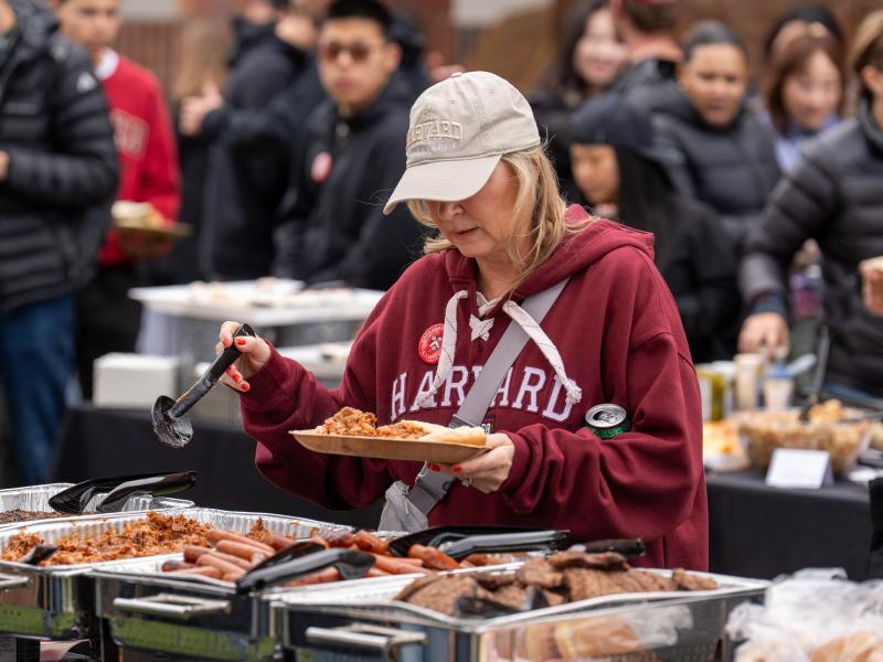 A woman in a Harvard hat and sweatshirt puts food on her plate in the buffet line.