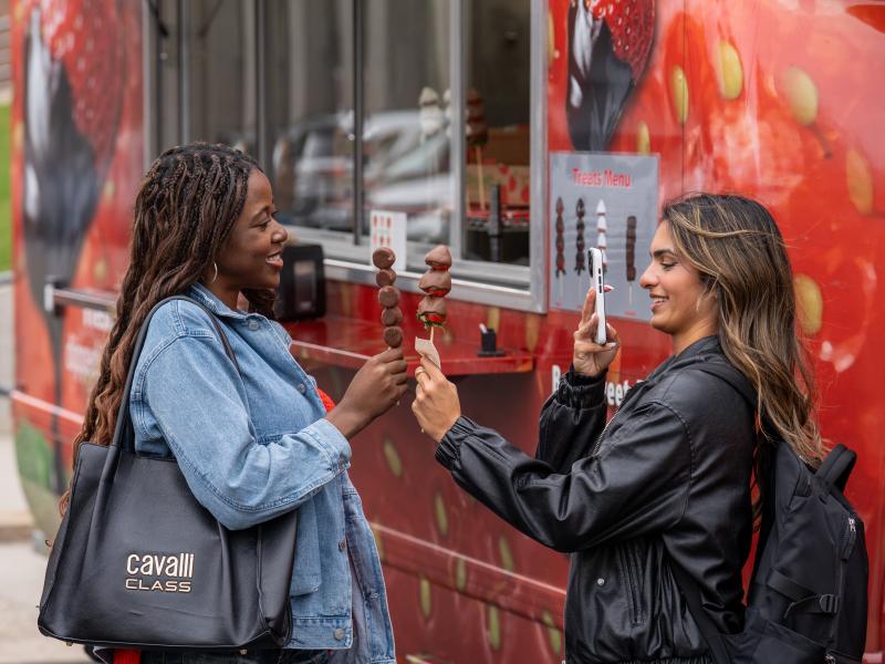 Two student girls take pictures of themselves holding their chocolate covered strawberries from a food truck.