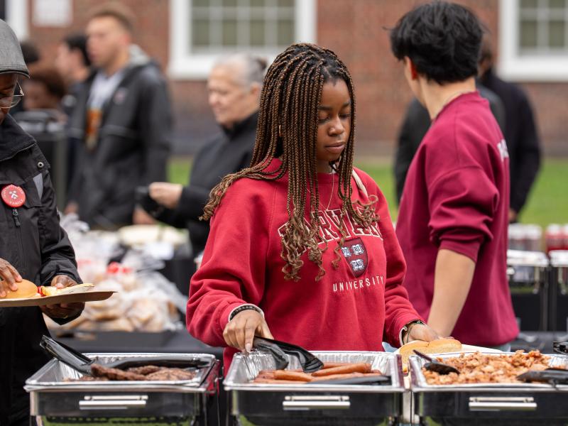A female student in a red Harvard sweatshirt picks up a hot dog while in line at the buffet station.