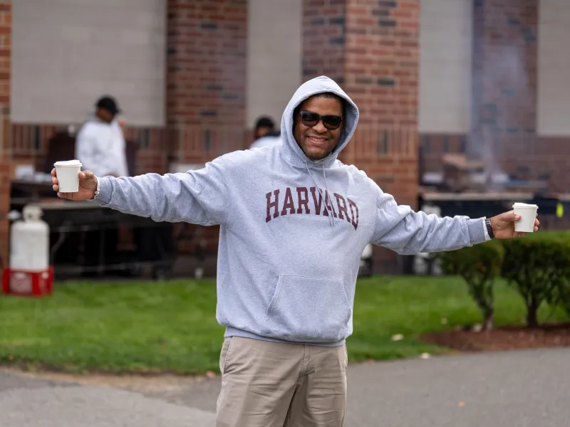 A man in a gray Harvard sweatshirt holds two white beverage cups in his outstretched hands and smiles.