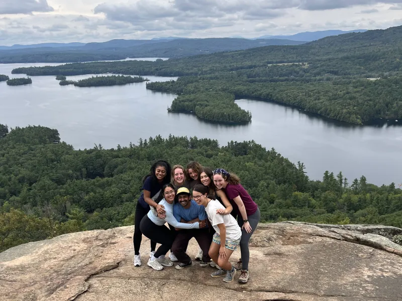 Lizbeth and seven peers posing on top of a mountain in New Hampshire, which a large lake in the background.