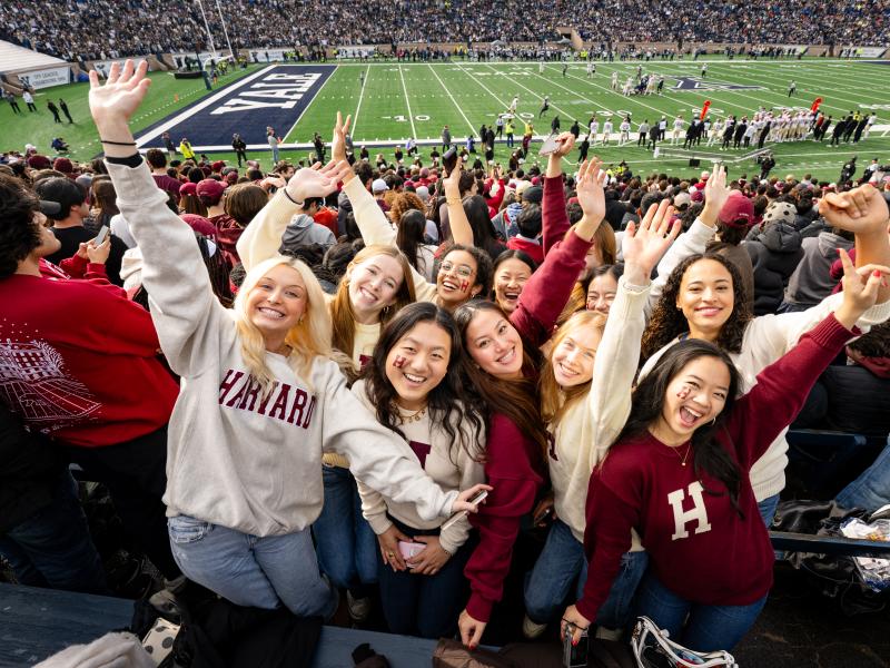 A group of young female Harvard College students stand together smiling with their arms outstretched and wearing Harvard sweatshirts.