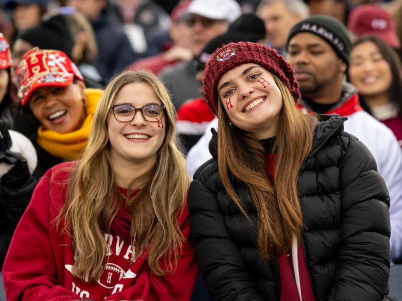 Two young female Harvard students smile together while wearing red Harvard merchandise.