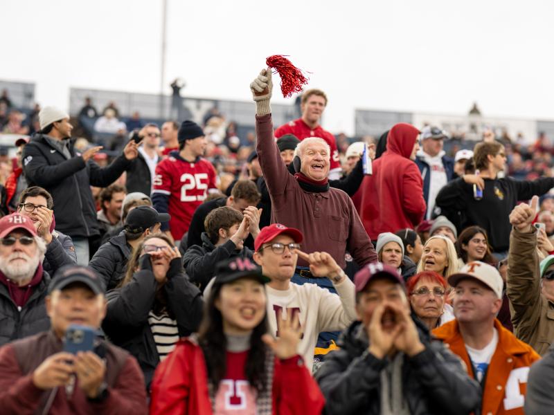 An older male stands above the crowd waving a red pom pom in the air to cheer for Harvard.