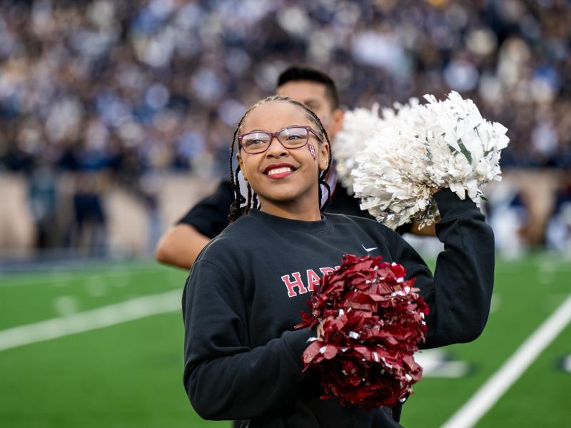A young black female Harvard cheerleader smiles and cheers with her red and white pom poms.