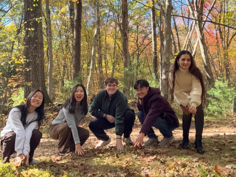 A group of students crouch and smile while touching the ground of Harvard Forest.