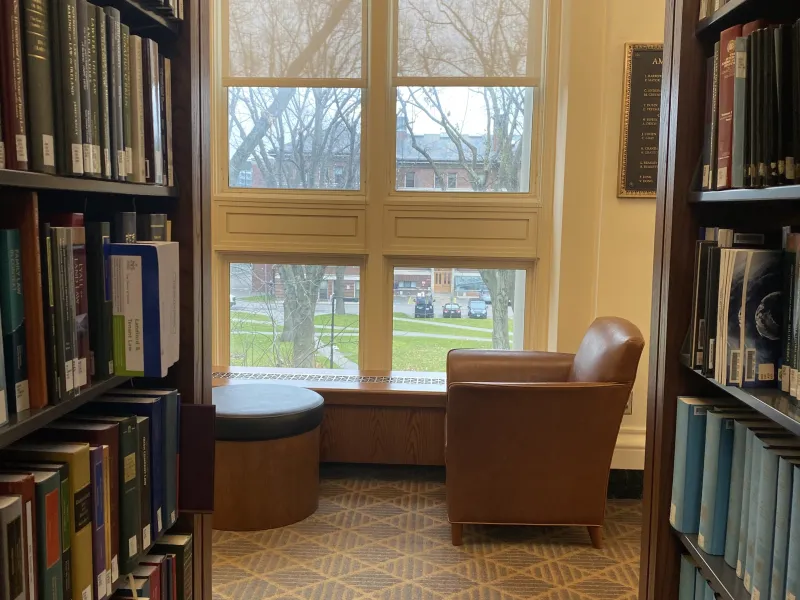 A singular chair located near rows of books on one side and a window view of Harvard Yard on the other.