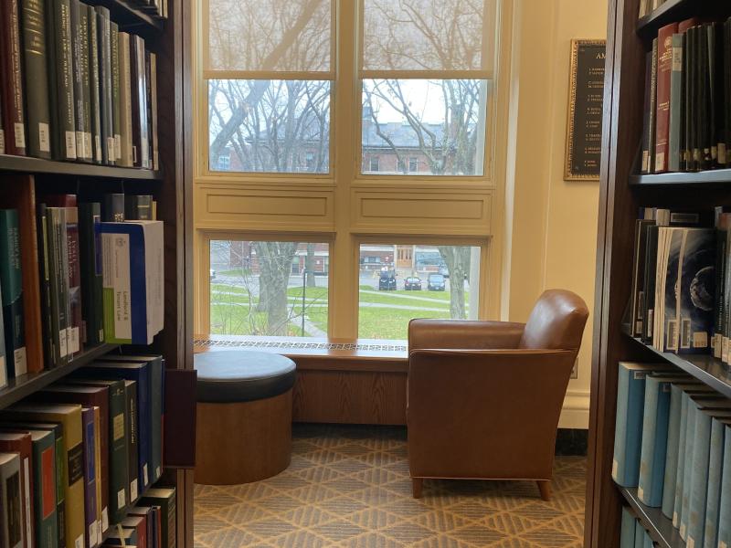 A singular chair located near rows of books on one side and a window view of Harvard Yard on the other.