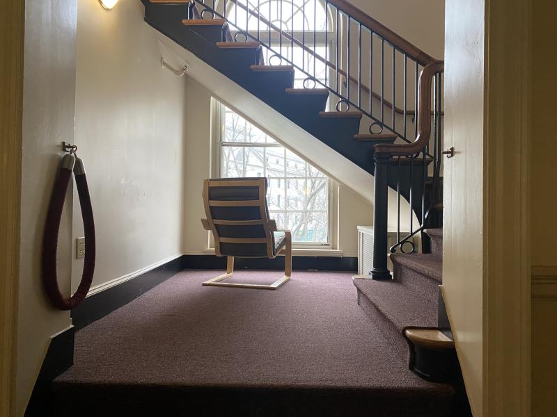 A lone chair seated next to a large window, underneath a staircase.