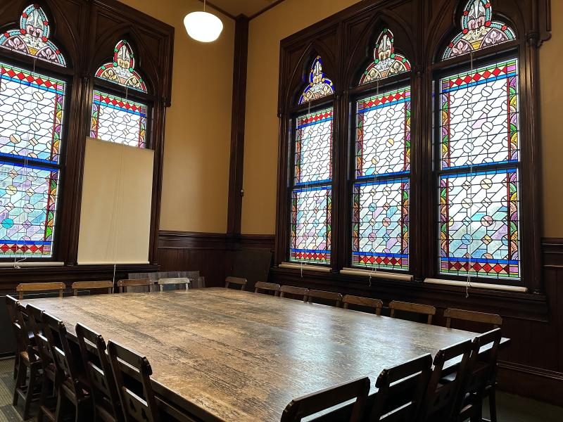 Stained glass windows surrounding a wooden table and chairs with a large white notepad in the corner.