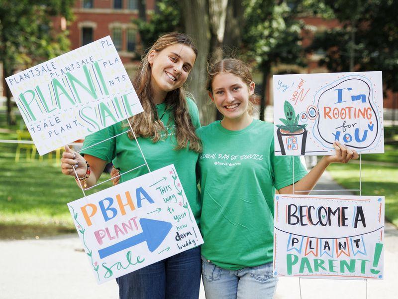 Two female students wearing green shirts and holding up handmade signs that promo the Plant Sale.
