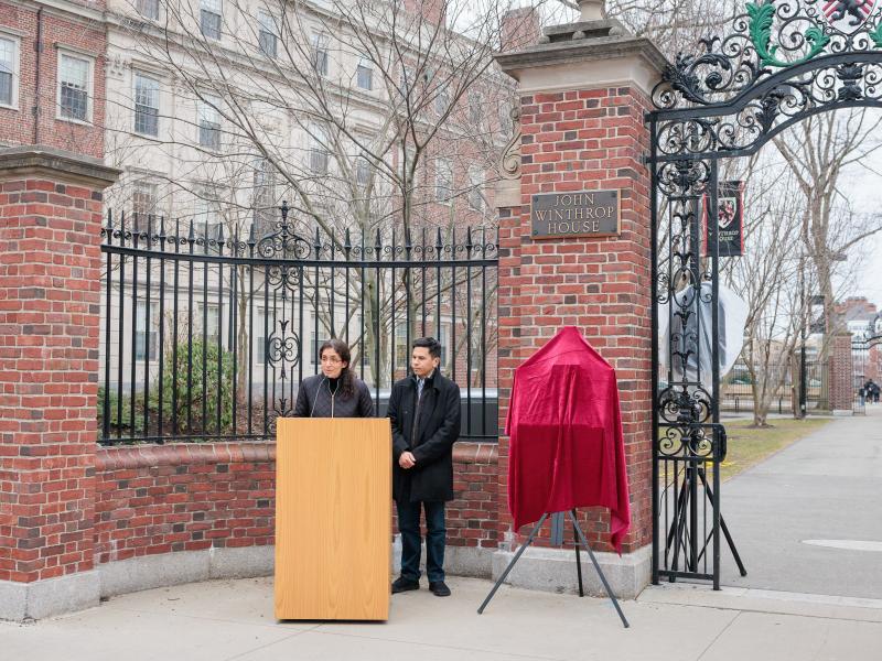 Two speakers stand at a podium by the John Winthrop House gate beside a plaque covered with a red cloth.