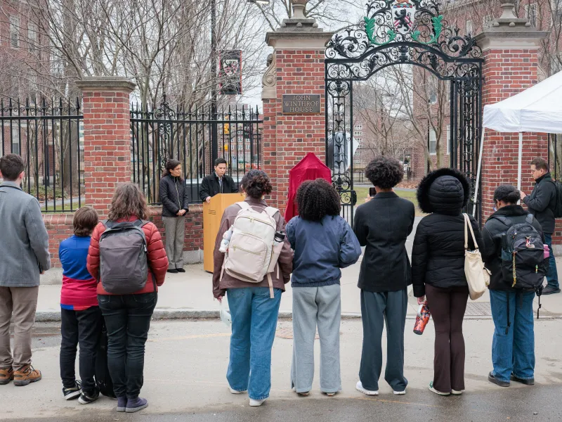 Audience stands facing speakers at a podium by the John Winthrop House gate and a plaque covered in red cloth.