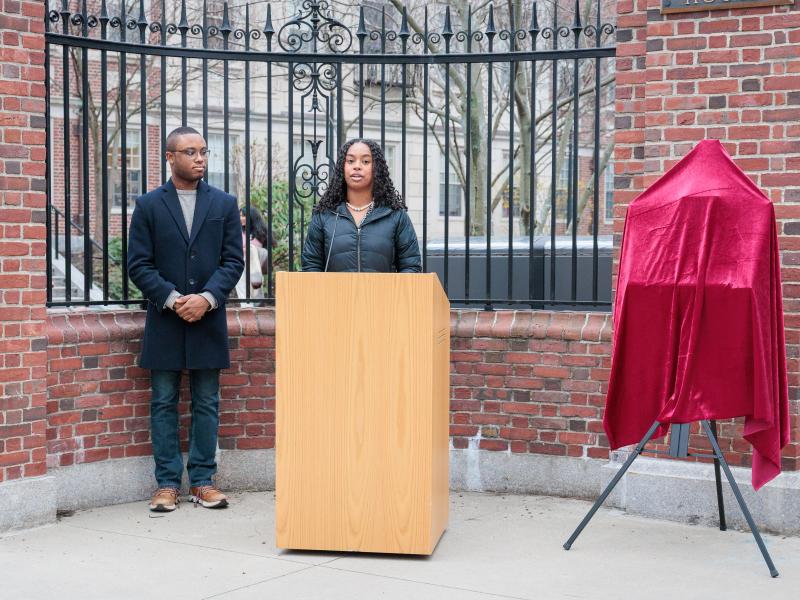 Two speakers stand at a podium near a brick gate, beside a plaque covered with a red cloth.