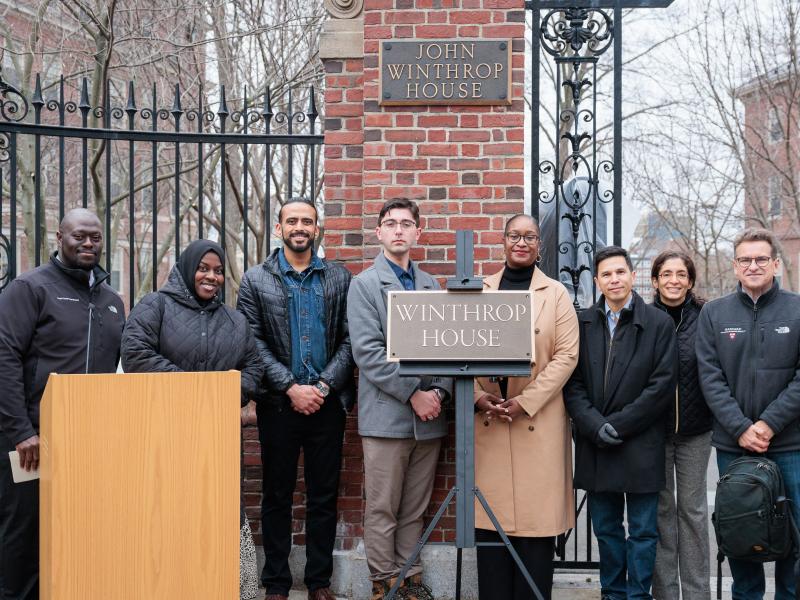 Group of people stand smiling by the Winthrop House gate and sign during an outdoor ceremony.