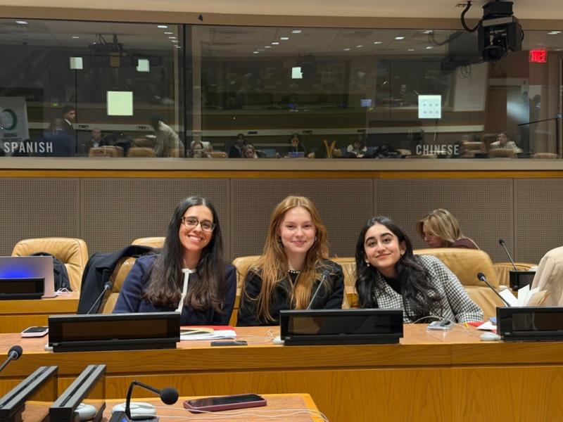Three young women sit smiling behind microphones at conference desks in a UN-style meeting room.